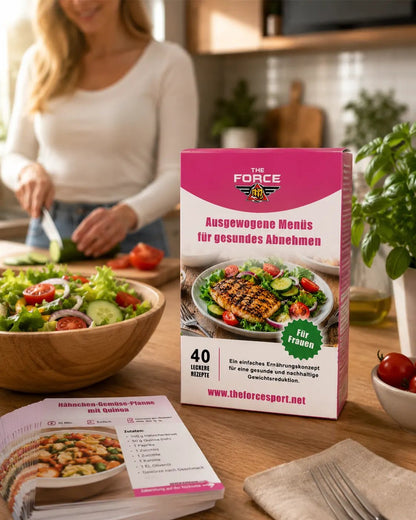 A pink and white box of 'The FORCE' healthy meal plan booklet stands on a wooden table in a kitchen setting, with a blurred woman preparing food in the background, a salad bowl, and an open recipe booklet in the foreground.