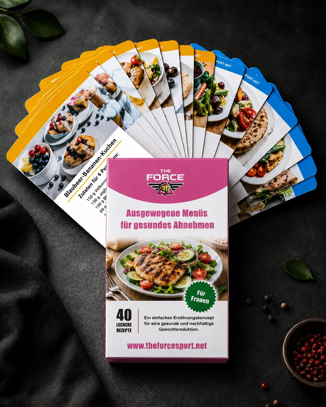 A boxed card deck titled "THE FORCE" with the German text "Ausgewogene Menüis für gesundes Abnehmen" is displayed in front of a fanned-out set of recipe cards on a dark textured surface. The box is pink and white with a salad photo, and the cards show colorful food and recipe imagery.